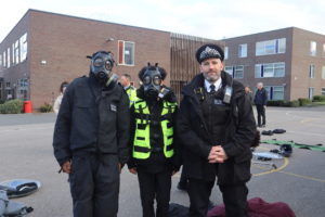 Two students stand in gas masks alongside a Police Officer