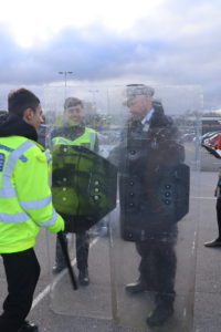 A uniformed police officer stands behind a riot shield whilst a student, wearing a high vis coat watches.